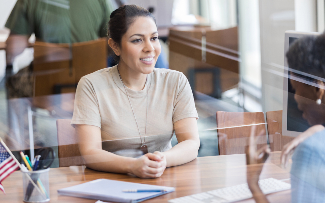 A cheerful female soldier smiles as a female bank employee discusses the bank's services; the bank employee gestures while talking with the soldier